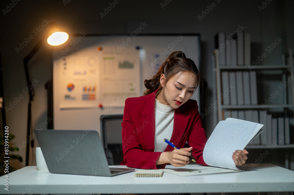 A woman in a red jacket is sitting at a desk with a laptop and a piece of paper. She is writing on the paper with a pen. The scene suggests a professional setting, possibly in an office or a workspace A woman in a red jacket is sitting at a desk with a laptop and a piece of paper. She is writing on the paper with a pen. The scene suggests a professional setting, possibly in an office or a workspace