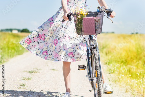 Woman in dress with flowers carries wild flowers bouquet on bicycle in basket , flowers  gathered in a meadow  by woman in the countryside in summer time sunny day