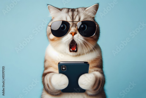 Shocked cat in sunglasses holding smartphone on color background