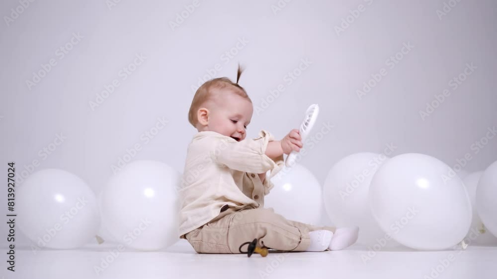 Little adorable Caucasian baby sits on the floor in studio among the white balloons. Cute child plays with toys and pulls it to mouth. Little adorable Caucasian baby sits on the floor in studio among the white balloons. Cute child plays with toys and pulls it to mouth.