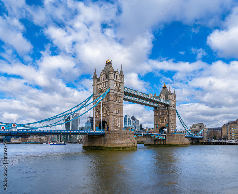 Obraz premium Panoramic view of London's Tower Bridge from the River Thames with maritime traffic and ferries sailing along the river under a blue sky with white clouds. United Kingdom.