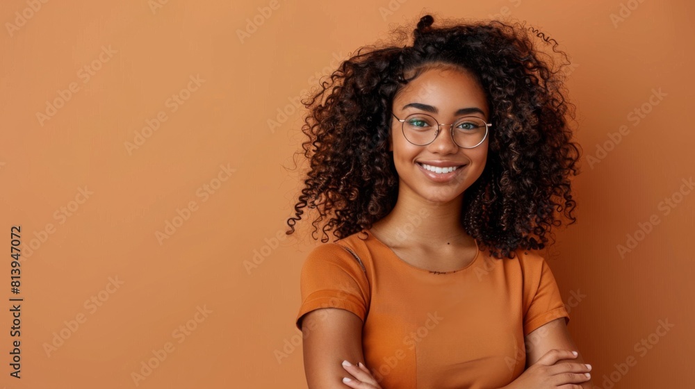 A cheerful young woman with curly hair and glasses posing against an orange background with a confident smile. A cheerful young woman with curly hair and glasses posing against an orange background with a confident smile.