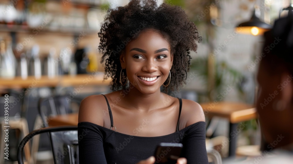 A smiling young African American woman holding a smartphone engaged in a lively conversation in a cafe setting. A smiling young African American woman holding a smartphone engaged in a lively conversation in a cafe setting.