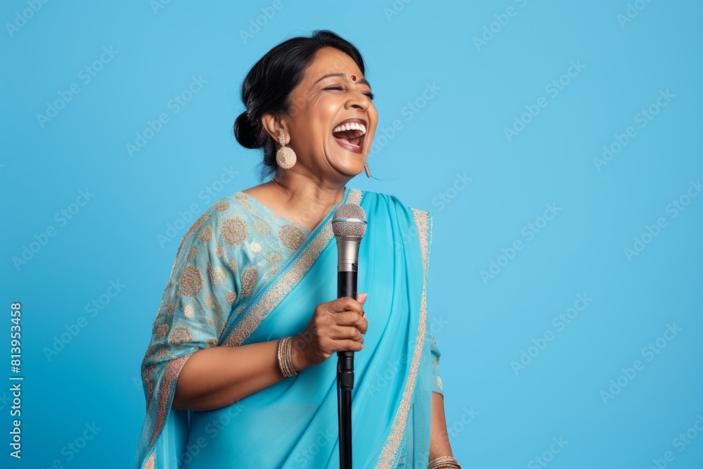 Portrait of a grinning indian woman in her 50s dancing and singing song in microphone isolated on pastel blue background Portrait of a grinning indian woman in her 50s dancing and singing song in microphone isolated on pastel blue background