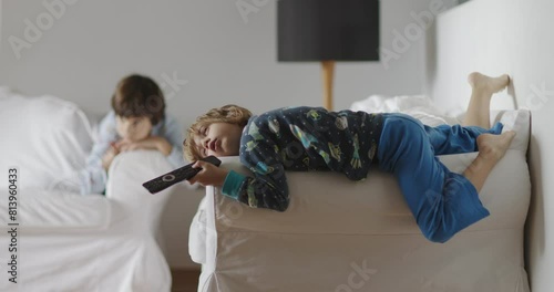 Two kids lounge idly in a living room, one lying on a couch with a remote in hand, eyes glued to the TV, the other sitting nearby, a picture of lethargy.