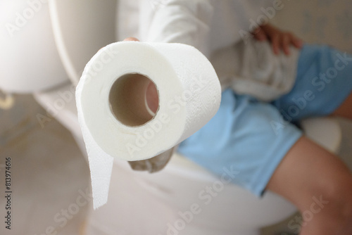Man sitting on the toilet bowl and his hand is holding toilet paper or tissue close-up over blurred background in bathroom for concept of excretion and hygiene.