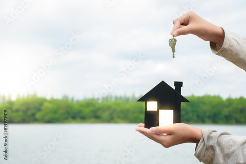Man hands holding miniature black house model building and holding keys with dot soft light on lake and green forest background over sky for concept of home insurance and real estate. photo