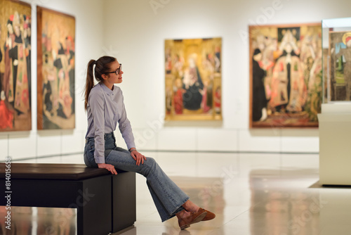 Thoughtful young Caucasian woman wearing glasses and looking at exhibition. Concept of Museum Day.