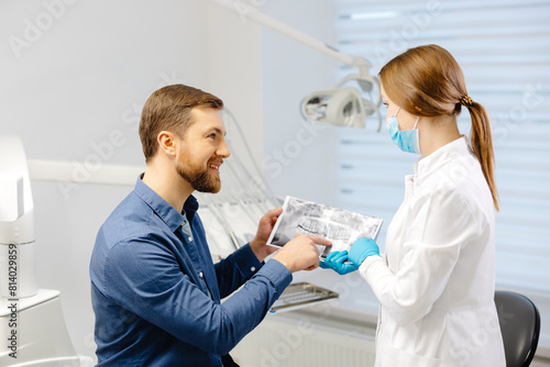 Young attractive man visiting dentist  sitting in dental chair at modern light clinic. Young woman dentist holding x ray image.