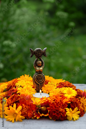 Triple Goddess figurine with candle and colorful flowers in garden, nature background. Symbol of the Triune Moon. esoteric ritual for Litha, Midsummer. witchcraft, wiccan spiritual practice.
