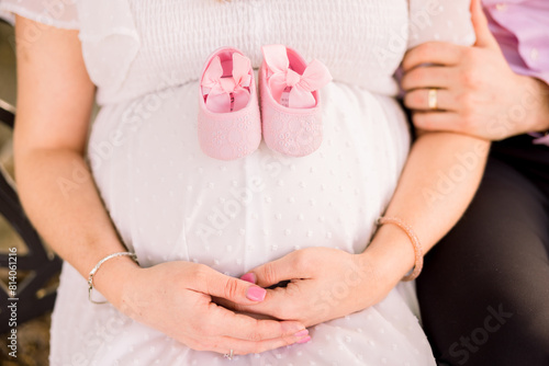  A married couple sits together, and she cradles her hands around her pregnant bump and has two pink baby booties on top of her stomach. 