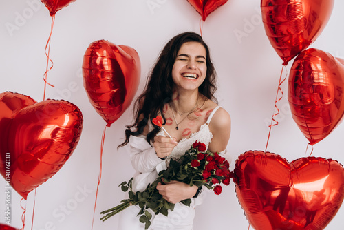 A smiling woman with kisses lipstick is holding a bouquet of red roses, candy, and standing amidst heart-shaped helium balloons, possibly celebrating a special occasion like Valentines Day
