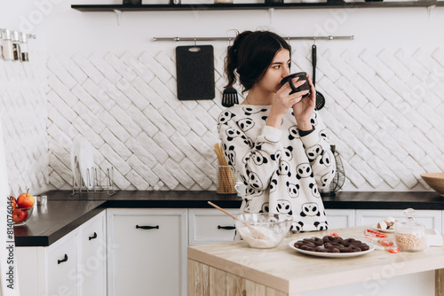 Cheerful woman standing in bright kitchen drinking hot beverage coffee tea, dressed in patterned pajama set. The kitchen counter arranged with ingredients, utensils, atmosphere domestic