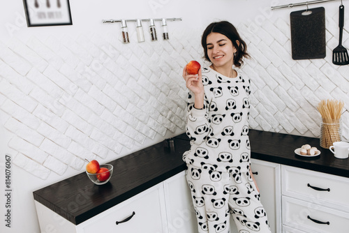 Cheerful woman standing in bright kitchen smiling laughing eating apple fruit, dressed in patterned pajama set. The kitchen counter arranged with ingredients, utensils, atmosphere domestic