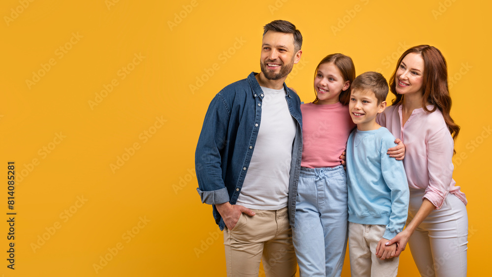 Fototapeta premium A happy family, with two parents and two children, embrace each other against a vibrant yellow backdrop. They appear cheerful and content, looking at copy space