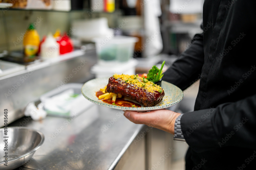A person holding a plate with a cooked steak and fries, garnished with herbs, in a kitchen setting