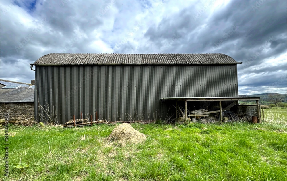 A large, industrial-looking metal barn, with a corrugated roof stands in the center of a green field under a cloudy sky high above, Silsden, Yorkshire, UK A large, industrial-looking metal barn, with a corrugated roof stands in the center of a green field under a cloudy sky high above, Silsden, Yorkshire, UK