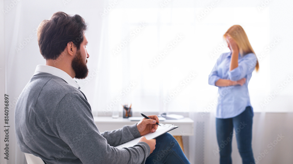 Obraz premium Man therapist sits in profile, attentively jotting down notes on a clipboard, listening to woman client who stands in the background with her hand on her forehead, suggesting distress.