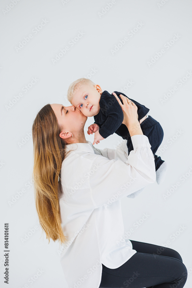 A young mother sits on a white background, holds her baby in her arms and kisses her. Mother and little son. A young mother sits on a white background, holds her baby in her arms and kisses her. Mother and little son.