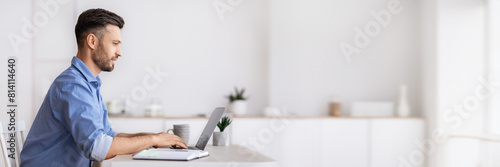 Handsome millennial businessman sitting at workplace in white office, working on laptop, typing on computer keyboard, answering emails, corresponding with clients, side view with copy space