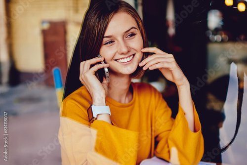 Positive young woman with short haircut laughing during phone call on smartphone sitting at laptop device and working remotely in coffee shop.Cheerful pretty hipster student talking on cellular
