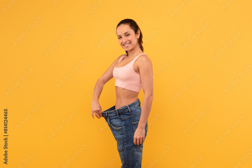 A cheerful young woman stands against a yellow backdrop, showing off her weight loss success by holding out the waistband of her oversized blue jeans A cheerful young woman stands against a yellow backdrop, showing off her weight loss success by holding out the waistband of her oversized blue jeans