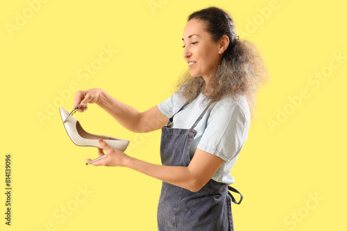 Female shoemaker measuring high-heeled shoe on yellow background