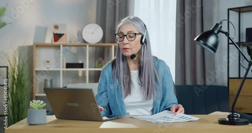 Senior woman in headphones holding online meeting on laptop computer with coworkers from home office. Distant work concept.