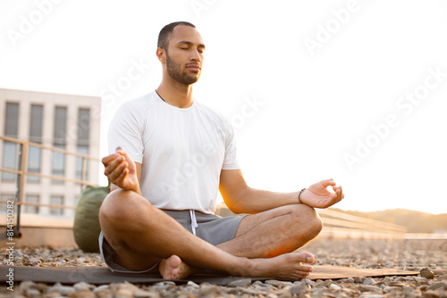 Handsome male meditating in lotus position while sitting on mat on roof of modern house. Young athletic man doing yoga at sunset on urban background.