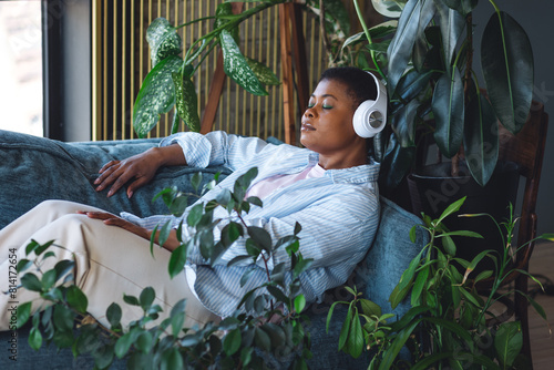 Concept of relaxation and meditation at home. Young plus size calm African American woman relaxing on the couch, listening to favourite music in headphones. Mindfulness, wellbeing, work life balance