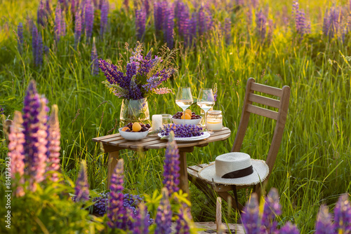 Private wedding party for two, table set with floral decor and greenery, fruits, wine in lupine purple field. Summer, sunset, golden hour. Romantic surprise, marriage proposal. Elegant decoration