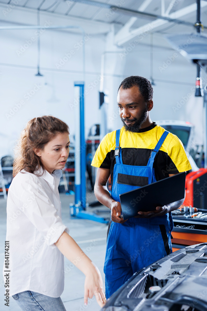 Obraz premium Smiling serviceman in car service uses tablet to calculate invoice after fixing broken automobile radiator. Specialist uses device to inform customer of final costs after mending vehicle