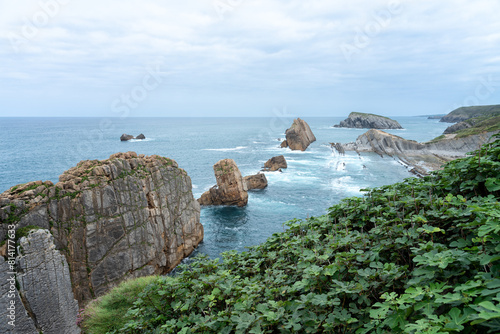 A rocky shoreline with a rocky cliff covered by green leafs and a rocky beach in the background. The ocean is calm and the sky is cloudy, Natural Park of the Dunes of Liencres, Cantabria, Spain.