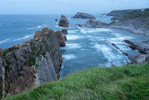 View from a cliff of a rocky beach with deep blue waters and green grass in the foreground, Costa Quebrada, Urros de Liencres, Cantabria, Spain.
