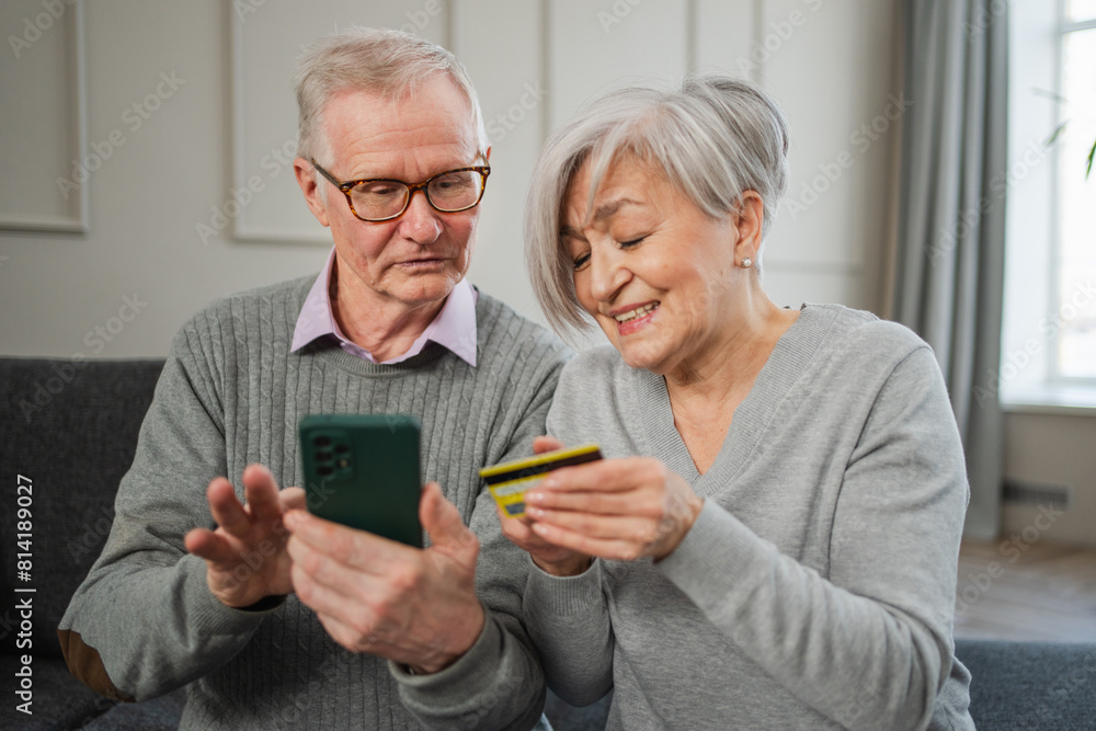 Senior couple man woman shopping online with smartphone paying with credit card. Old people buying on Internet enter credit card details at home indoors. Online shopping delivery service ecommerce Senior couple man woman shopping online with smartphone paying with credit card. Old people buying on Internet enter credit card details at home indoors. Online shopping delivery service ecommerce