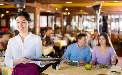 Portrait of positive Asian woman waitress standing in restaurant with dish.