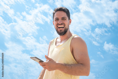 Young adult Laughing on beach with cell phone looking camera and sunny blue sky