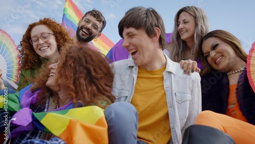 LGBT diverse group of friends laughing happy sitting together outdoor celebrating gay pride day. Joyful people from generation z community enjoying parade. Young liberals with rainbow flags and fans photo
