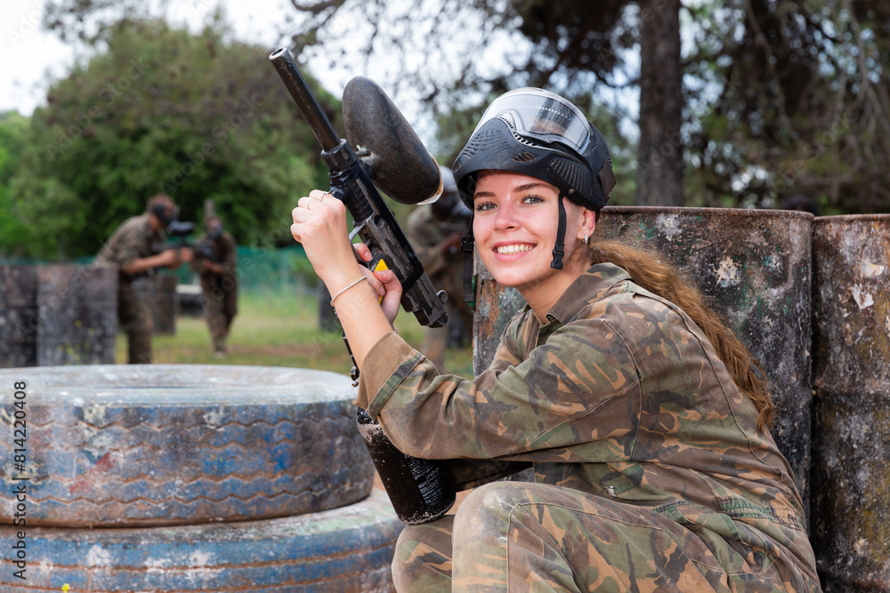 Fototapeta premium Young smiling woman paintball player in camouflage posing with gun outdoors