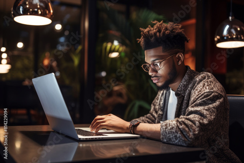 A black student using laptop and taking notes during an online class at a cozy coffee shop table during nighttime