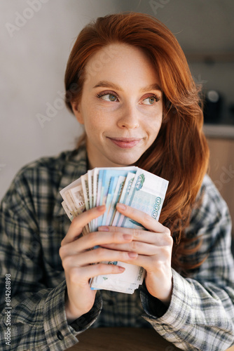 Vertical portrait of happy female freelancer holding in hands wad of Russian rubbles that she earned while working remotely at home. Remote earnings, online work. Young happy woman counting money.