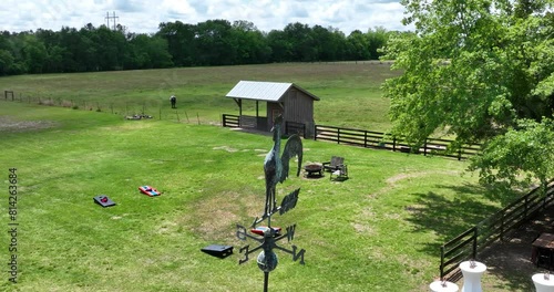 Closeup slow motion shot of a rooster wind vane on top of a tin roof barn with a game of cornhole set up on the ground below next to the pasture on a farm