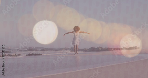 Image of lens flares over happy biracial young woman with arms outstretched walking at shore