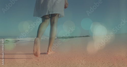 Image of lens flare over low section of biracial young woman walking barefoot on sandy beach