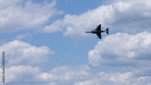 A military jet flies through a clear blue sky with minimal cloud cover.