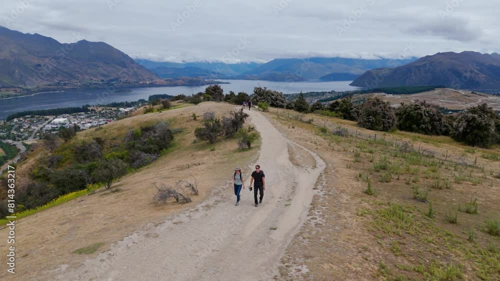 aerial - a couple hiking on a dirt trail in a hilly, scenic landscape near Wanaka, New Zealand, offering a panoramic view of the lake and surrounding mountains. aerial - a couple hiking on a dirt trail in a hilly, scenic landscape near Wanaka, New Zealand, offering a panoramic view of the lake and surrounding mountains.