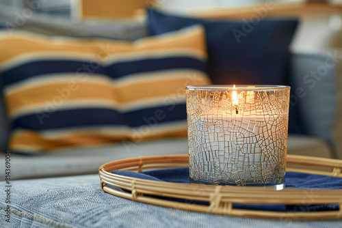A crackled glass candle jar on a bamboo tray table, with a navy and mustard striped pillow on a casual denim-covered sofa in the background.