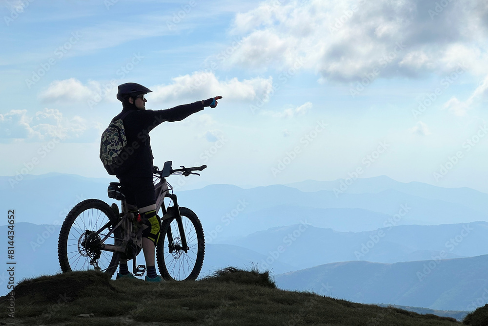 Cyclist man riding electric bike outdoors. Silhouette of male tourist resting on top of hill, pointing on beautiful mountain landscape, wearing helmet and backpack. Concept of active leisure. Cyclist man riding electric bike outdoors. Silhouette of male tourist resting on top of hill, pointing on beautiful mountain landscape, wearing helmet and backpack. Concept of active leisure.