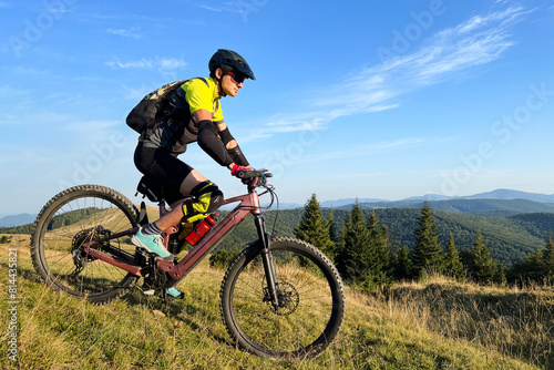 Cyclist man riding electric mountain bike outdoors. Male tourist biking along grassy trail in the mountains, wearing helmet and backpack. Concept of sport, active leisure and nature.