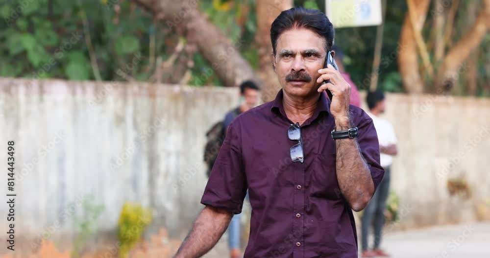 Indian man in a dark maroon shirt is talking on the phone on the street Indian man in a dark maroon shirt is talking on the phone on the street